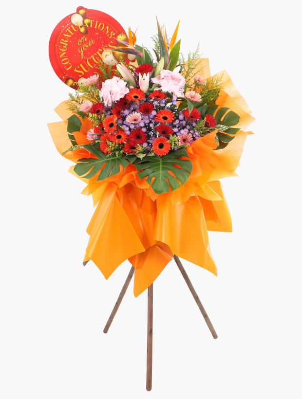A congratulatory flower arrangement adorned with an orange floral wrapper, elegantly displayed on a chic tripod stand, and featuring a red circular cardboard with the message "Congratulations on your success" affixed to it.